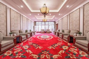 Una habitación de hotel con alfombra roja, dos sofás, una araña y un ventanal grande.