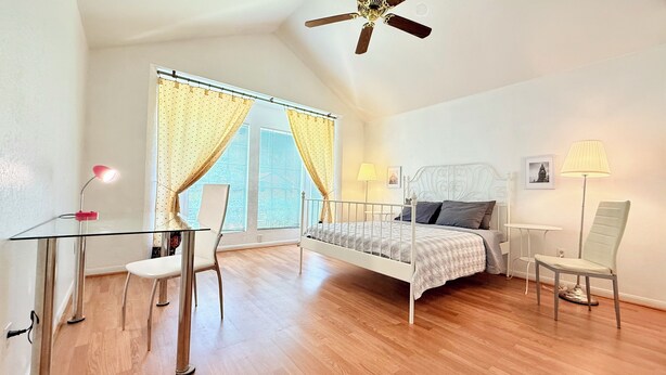 Une chambre à coucher comprenant un lit, un bureau, une chaise, un ventilateur de plafond et une fenêtre avec des rideaux.
