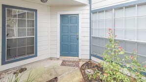 Une maison avec une porte bleue, une fenêtre avec des stores et un petit jardin agrémenté de plantes et d’un paillasson.