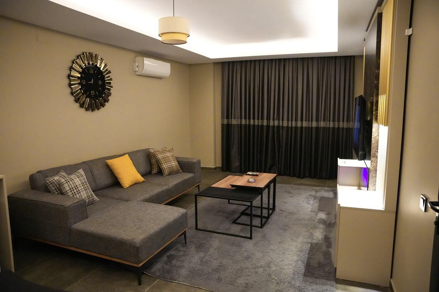 Una sala moderna con un sofá gris, una mesa de centro, un reloj de pared y un ventanal con cortinas.