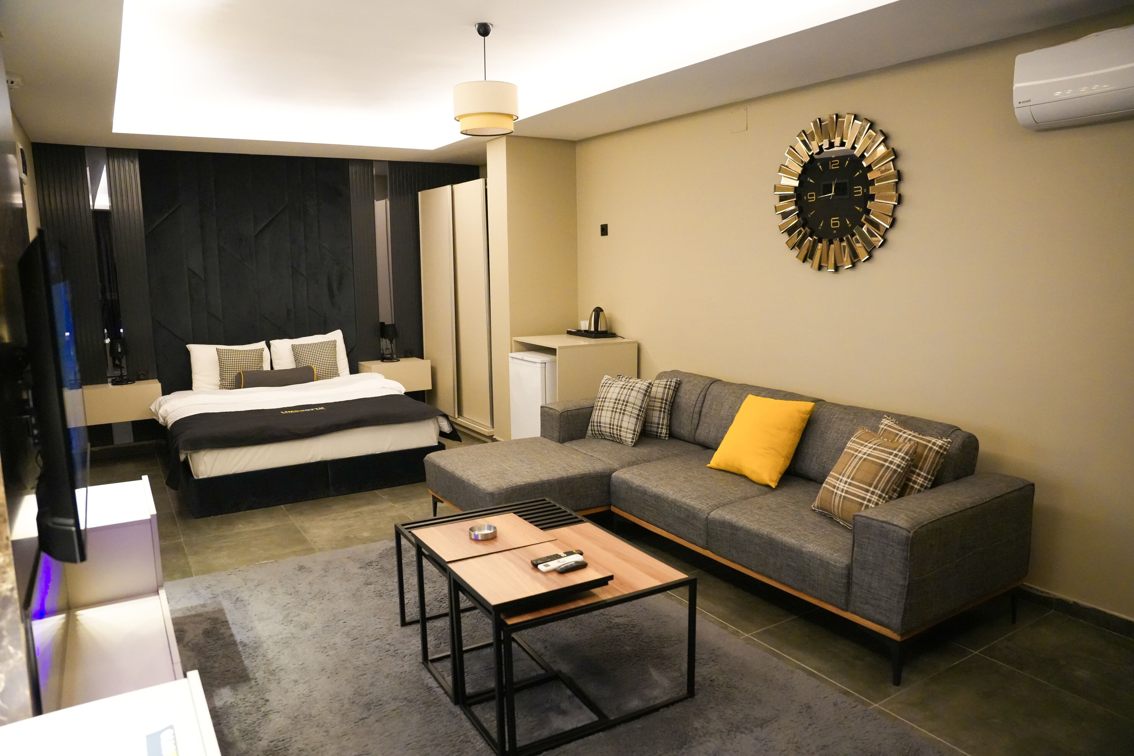 A modern hotel room with a bed, a gray sofa with yellow and plaid cushions, a coffee table, and a wall clock.