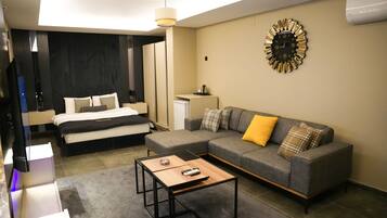 A modern hotel room with a bed, a gray sofa with yellow and plaid cushions, a coffee table, and a wall clock.