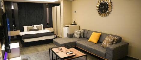 Una habitación de hotel moderna con una cama, un sofá gris con cojines amarillos y escoceses, una mesita de café y un reloj de pared.