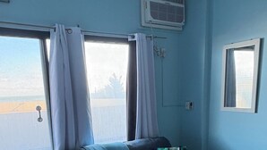 A room with a dark grey sofa, a blue throw blanket, and a floral pillow. There is a window with white curtains, a small air conditioning unit on the wall, and a tiled floor. - Oceanfront High Point North Studio M8 Just Steps to the Beach! (Ocean City)
