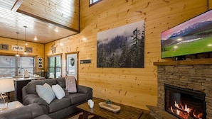 A cozy cabin interior with a stone fireplace, a television mounted above it, and a large forest-themed painting on the wall. - Charming Pet Friendly 3 Bedroom Chalet in Pigeon Forge with heated pool (Pigeon Forge)