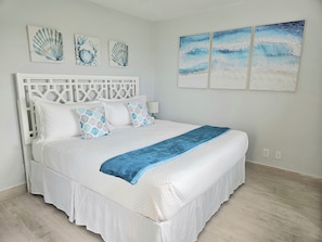 Quarto com cama branca, cabeceira branca e três quadros pendurados na parede.