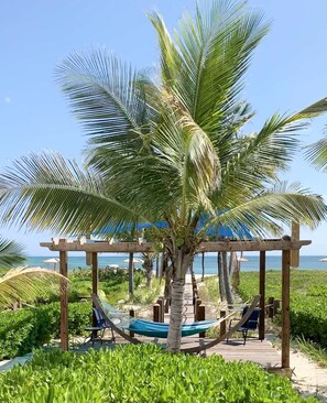 Uma pérgola de madeira com uma rede e um banco, cercada por vegetação exuberante e uma vista ampla do oceano.