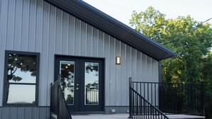 A building with a grey exterior, a black door, and a wooden staircase leading to a porch with a black railing. - On Beaver Lake! Rogers, AR, NEW Spacious Barndominium With Sweeping Lake Views. (Rogers)