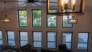 A spacious living room with multiple windows, a ceiling fan, and hanging light fixtures. - On Beaver Lake! Rogers, AR, NEW Spacious Barndominium With Sweeping Lake Views. (Rogers)