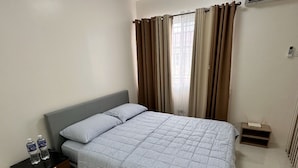 A neatly arranged bedroom with a bed, a bedside table, a nightstand, a window with curtains, and an air conditioning unit.