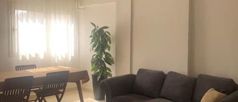 Un salón con un sofá gris oscuro, una mesa de comedor de madera con sillas, una planta en maceta y un candelabro en el techo.