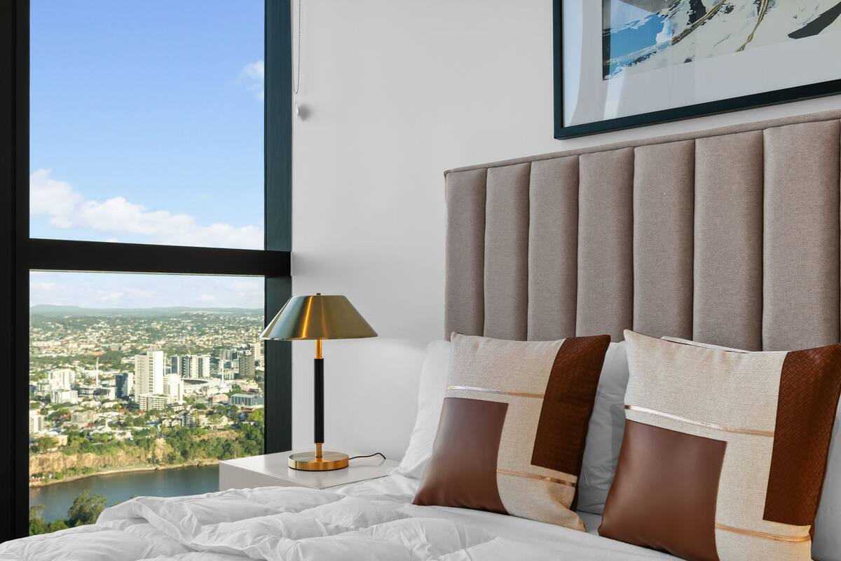 Ein Schlafzimmer mit großem Fenster, einem Bett mit weißer Bettwäsche, einer modernen Lampe und Blick auf die Stadt.