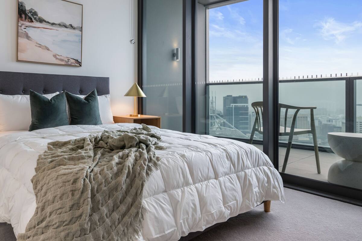 Ein modernes Schlafzimmer mit einem großen Bett, zwei Nachttischen, einem Beistelltisch, einem Gemälde und einem Balkon mit Blick auf die Stadt.