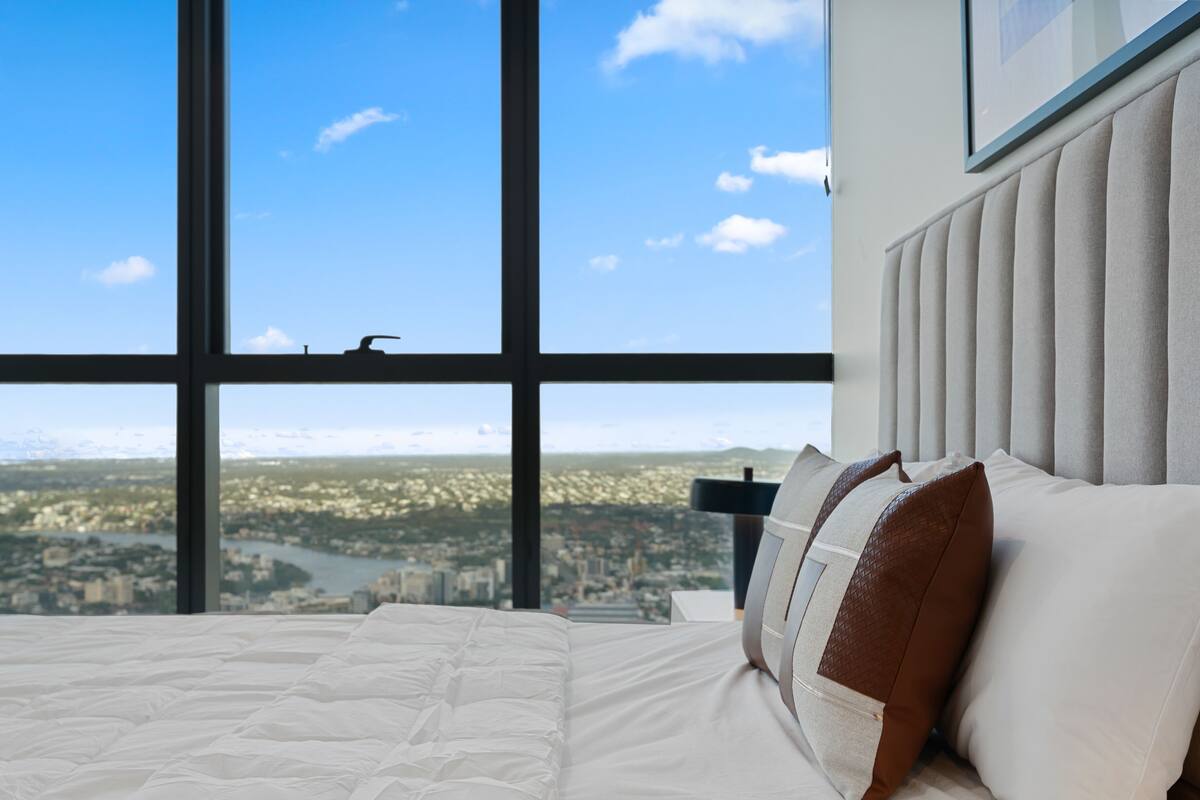 Ein Schlafzimmer mit großem Fenster, einem Bett mit weißer Bettwäsche und Blick auf eine Stadtlandschaft.