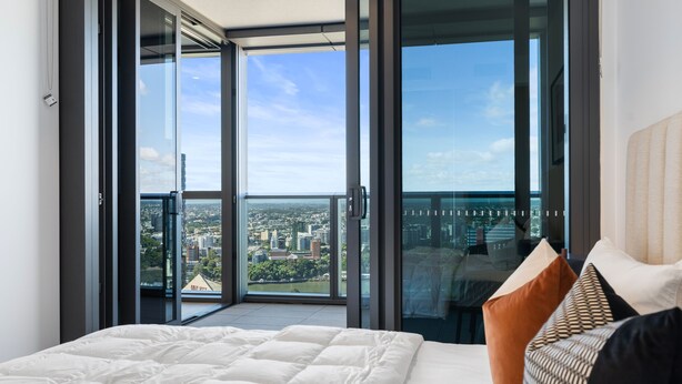 A modern bedroom with a large bed, a city view through floor-to-ceiling windows, and a balcony. - Facehome QWR (Brisbane)