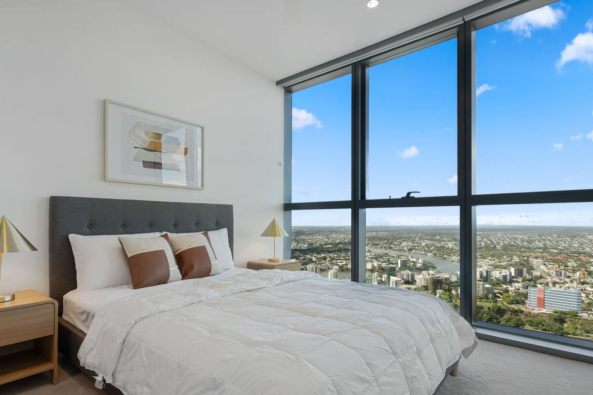 Ein Schlafzimmer mit großem Fenster, einem Bett mit weißer Bettwäsche, zwei Nachttischen und einem gerahmten Kunstwerk an der Wand.