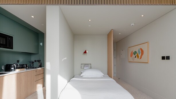 A modern hotel room with a kitchenette, a bed with white bedding, a framed abstract painting, and a small red lamp.