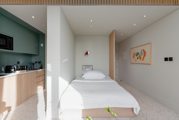 A modern hotel room with a kitchenette, a bed with white bedding, a framed abstract painting, and a small red lamp.