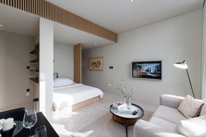 A modern hotel room with a bed, a round coffee table with a vase and remote control, a sofa, and a TV on the wall.