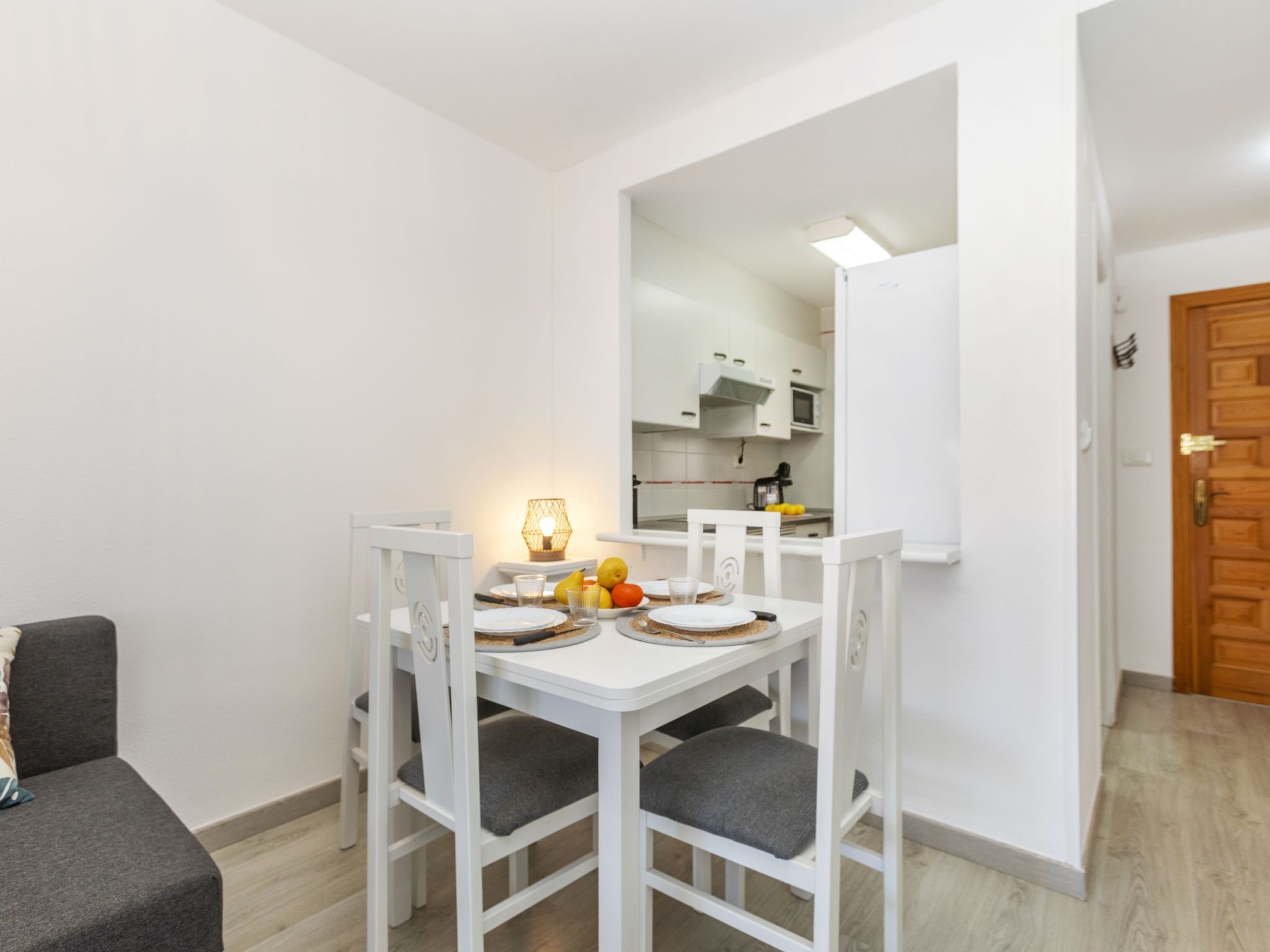 Een gezellig eetgedeelte met een witte tafel en stoelen, een klein keukentje zichtbaar door een open doorgang, en een grijze bank op de voorgrond.
