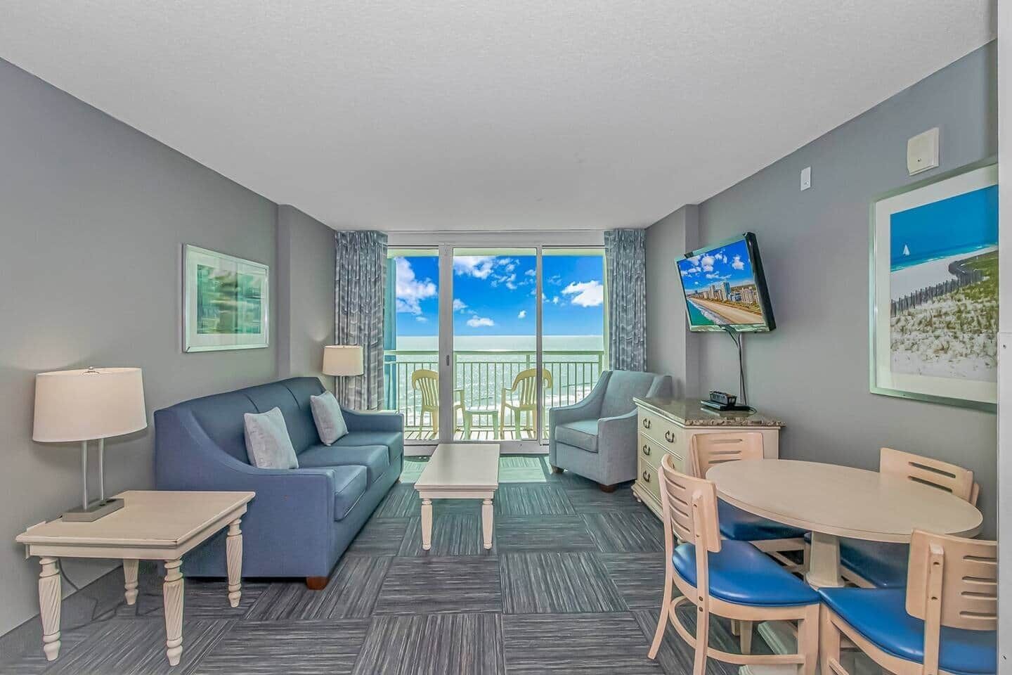 A hotel room with a balcony, a sofa, a dining table, and a TV.