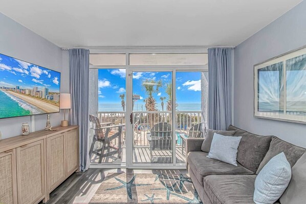 A balcony with a view of the beach and ocean, a sofa, a TV, and a dining area.