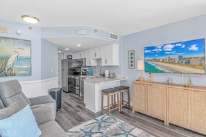 A modern kitchen with white cabinets, a central island, and a dining area with a gray sofa and a star-patterned rug. - Luxe Oceanfront @ Seawatch Resort! (Horry County)