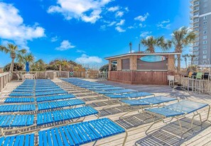 A sun deck with blue lounge chairs, a wooden structure with a sign, and a tall building in the background. - Luxe Oceanfront @ Seawatch Resort! (Horry County)