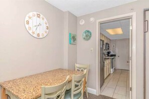 A dining area with a granite countertop, two chairs, a wall clock, and a kitchen visible through an open door.