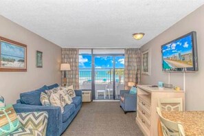 A room with a sofa, a desk, a chair, a TV, and a balcony with a view.