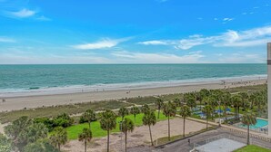 Vista a la playa con zona de piscina, palmeras y cielo despejado.