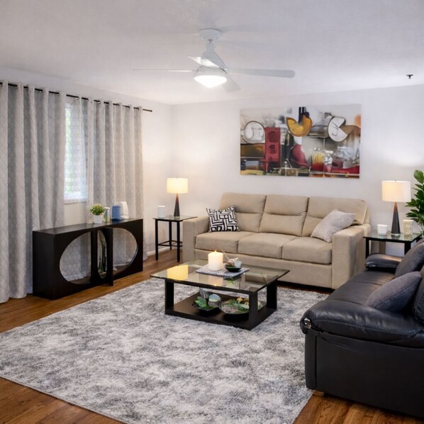 A living room with a beige sofa, a black leather chair, a glass coffee table, a black console table, and a painting on the wall.
