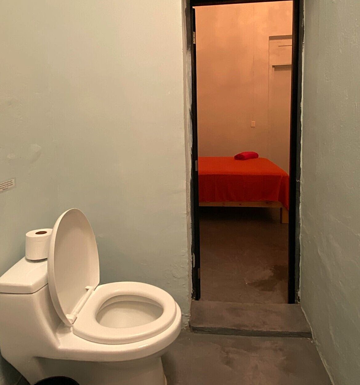A bathroom with a toilet, a roll of toilet paper, and a bed with a red blanket in the adjacent room.