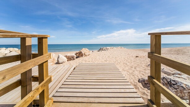 A wooden boardwalk leading to a sandy beach with large rocks and a clear blue sky.