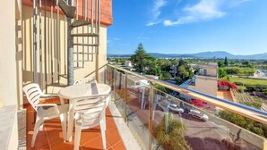 A balcony with a table and chairs, a glass railing, and a view of a cityscape and mountains.