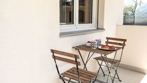 A small outdoor seating area with a table and two wooden chairs, a window, and a balcony with a view of trees.
