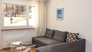 A modern living room with a grey sofa, a glass coffee table, and a view of a rustic outdoor area through a large window.