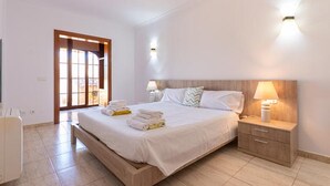 A bedroom with a bed, bedside tables, a nightstand, a door with a view, and a tiled floor. - Nice home in Maria de la Salut (Maria de la Salut)