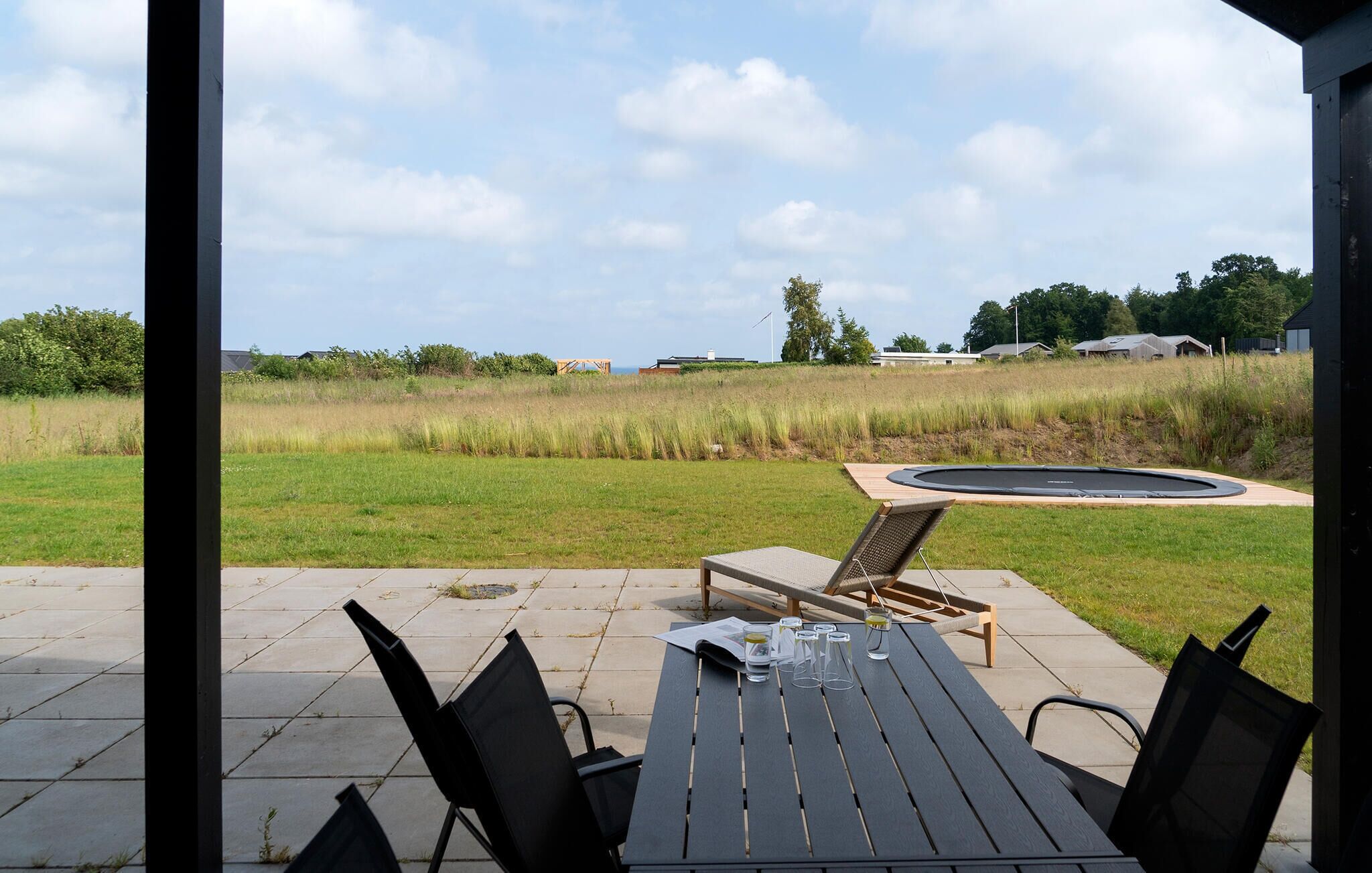 An outdoor patio with a table and chairs, a trampoline, and a view of a grassy field and houses in the distance.