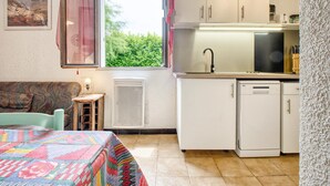 A compact kitchen with a window, a radiator, a small table with a lamp, and a colorful quilt on the bed.
