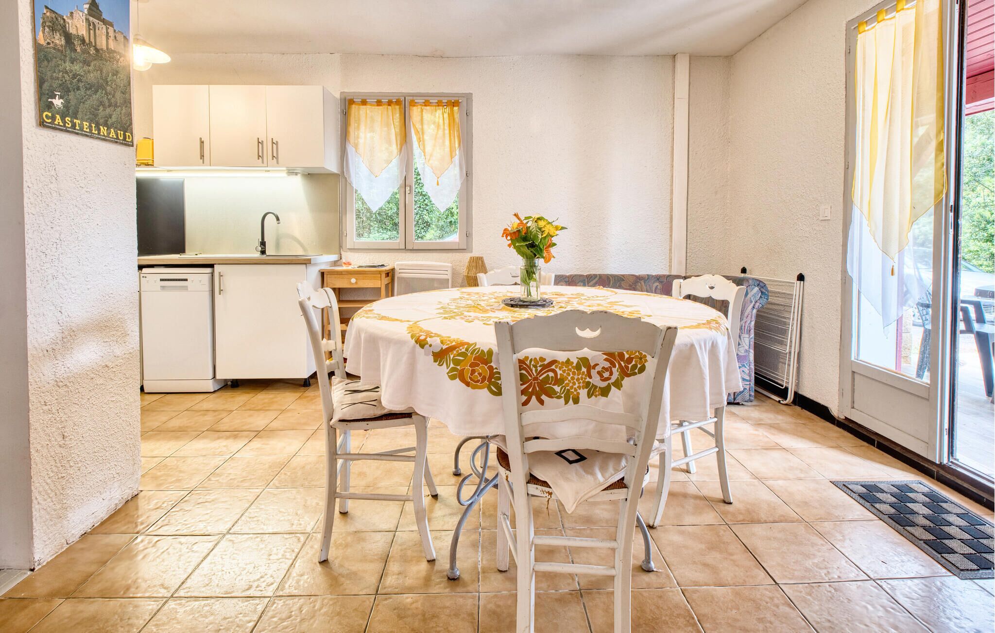 A compact kitchen with a sink, cabinets, and a window with curtains. There is a dining area with a table, chairs, and a vase of flowers.