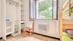 Una habitación con literas, una ventana que da a vista de árboles, un radiador, una silla y un armario blanco con estantes.