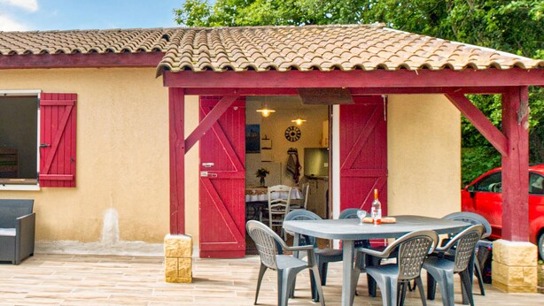 Un patio al aire libre con una mesa redonda y sillas, una pérgola roja y un coche rojo aparcado cerca.