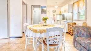 Un comedor con una mesa redonda, sillas blancas y un florero. Se observa una cocina al fondo con armarios y un reloj.