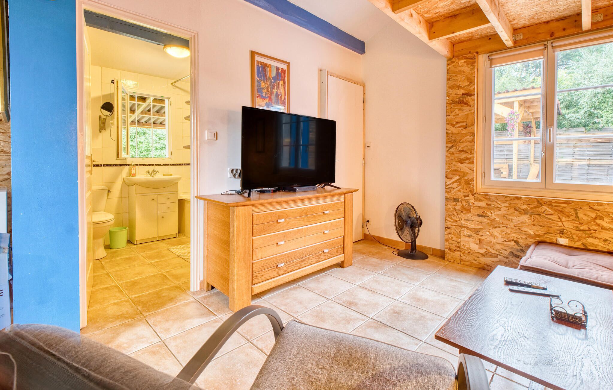 A room with a TV on a wooden dresser, a fan, a table with a remote and glasses, and a view of the outside through a window.