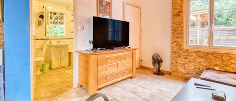 A room with a TV on a wooden dresser, a fan, a table with a remote and glasses, and a view of the outside through a window.