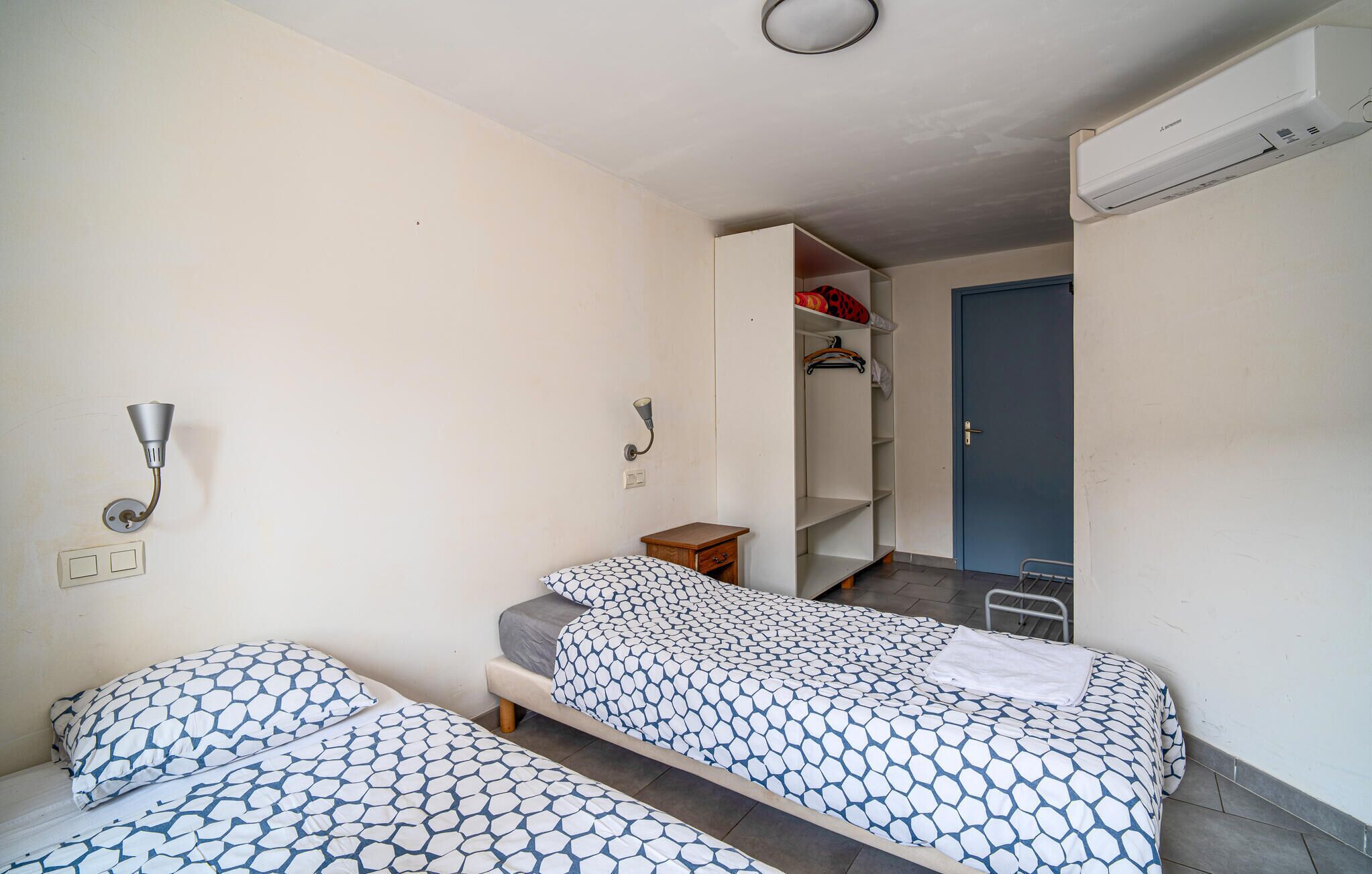 A bedroom with two beds, a wardrobe, a small bedside table, and a door leading to a bathroom.