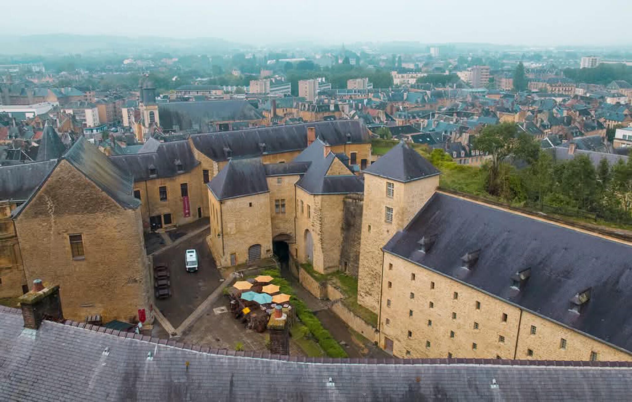 A historic building complex with multiple stone buildings, a courtyard with tables and umbrellas, and a view of the surrounding cityscape.
