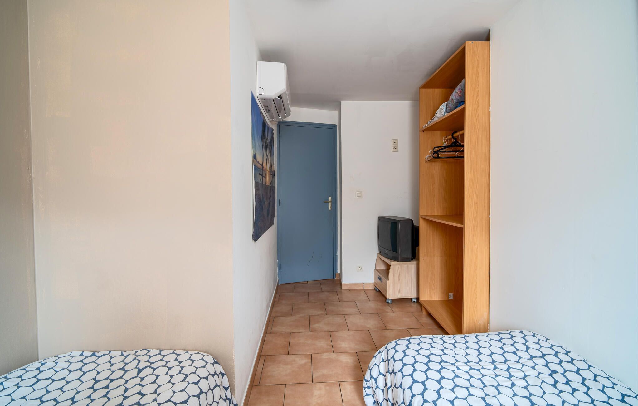 A small, narrow room with two beds, a wooden wardrobe, a television, and a door leading to another room.