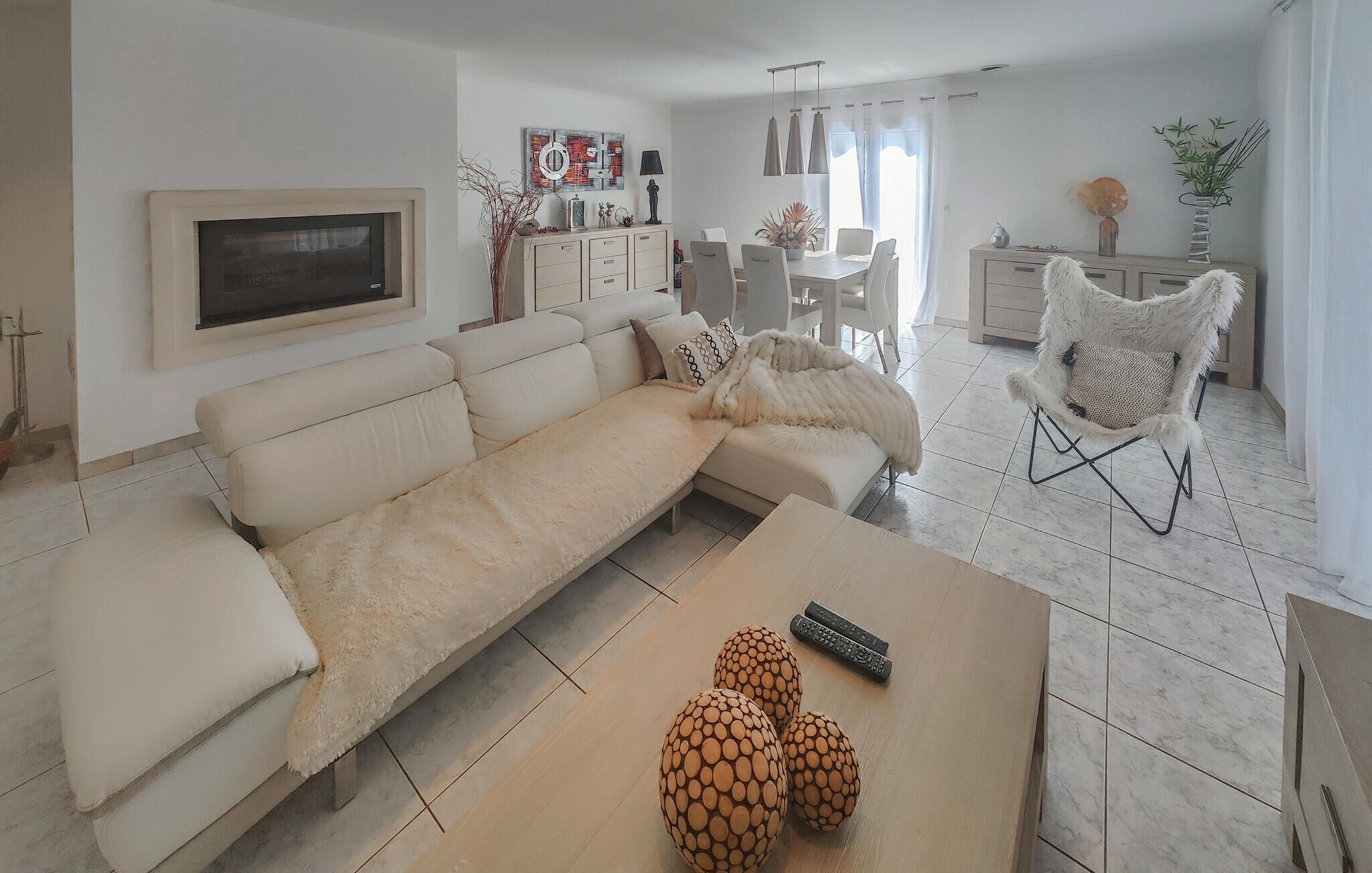 Una sala de estar moderna con un sofá color beige, una mesa de centro con objetos decorativos, y un comedor con una mesa y sillas blancas.